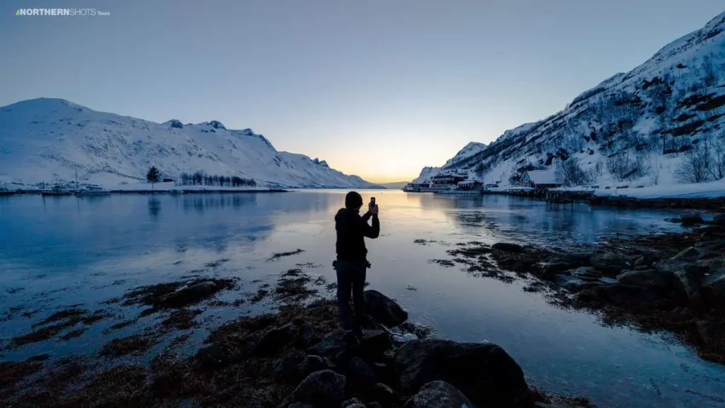 Visitor standing on stones at water’s edge, photographing the fjord at sunset with a mobile phone.