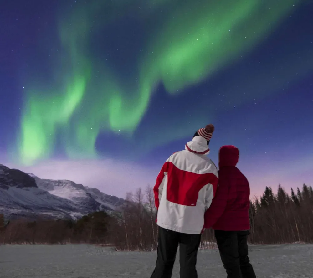 Rear view of a couple standing before snowy mountains and forest, watching the Northern Lights sweep across the night sky.