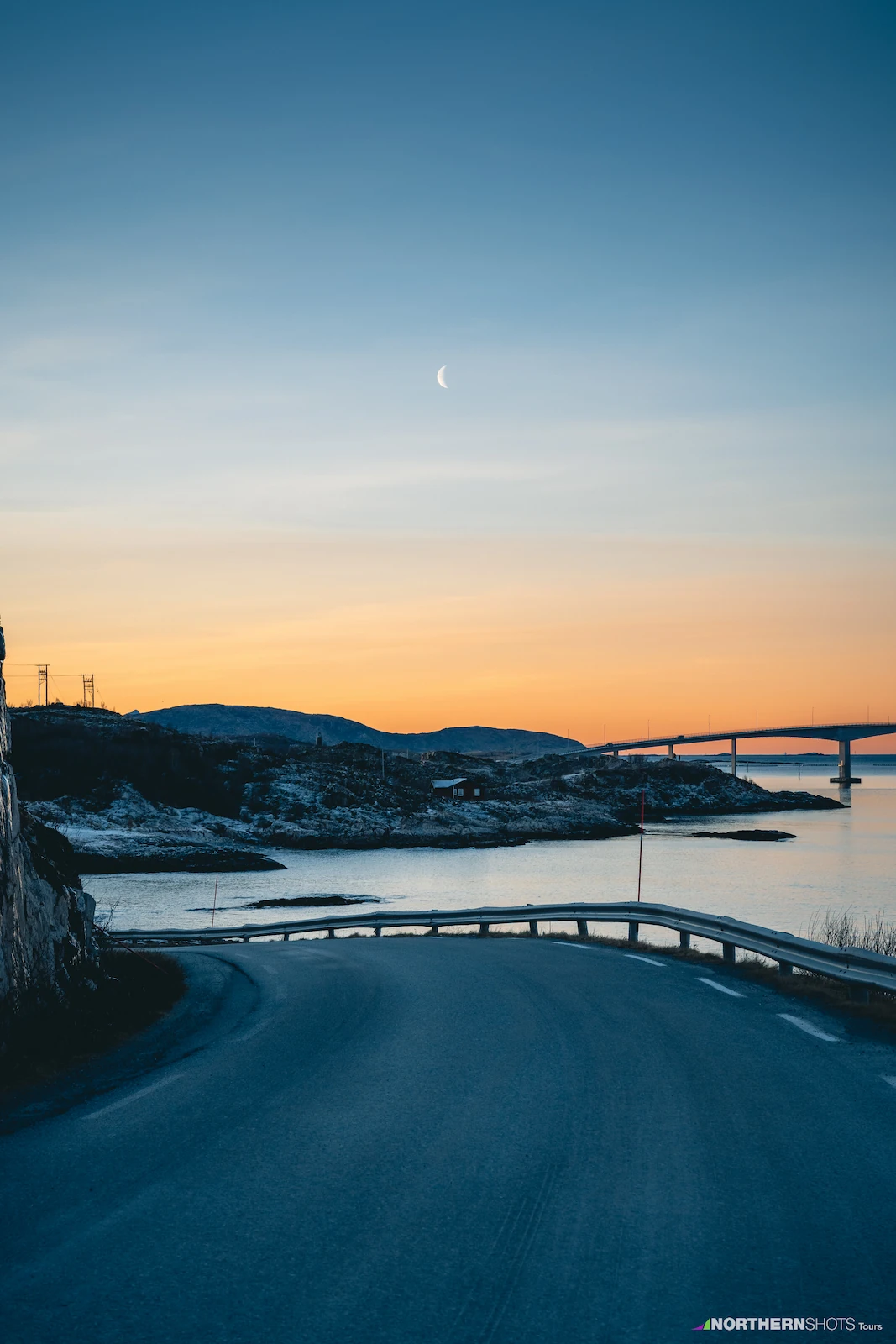 View approaching Sommarøy Bridge at dusk under an orange sky with a visible moon.